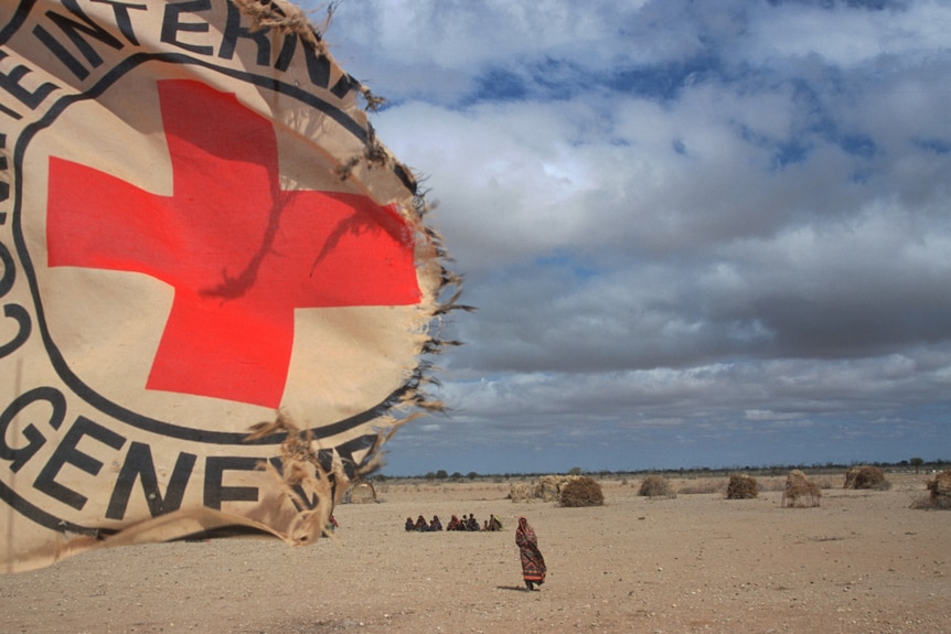 A ripped ICRC flag flaps in the wind in front of an arid landscape with refugees