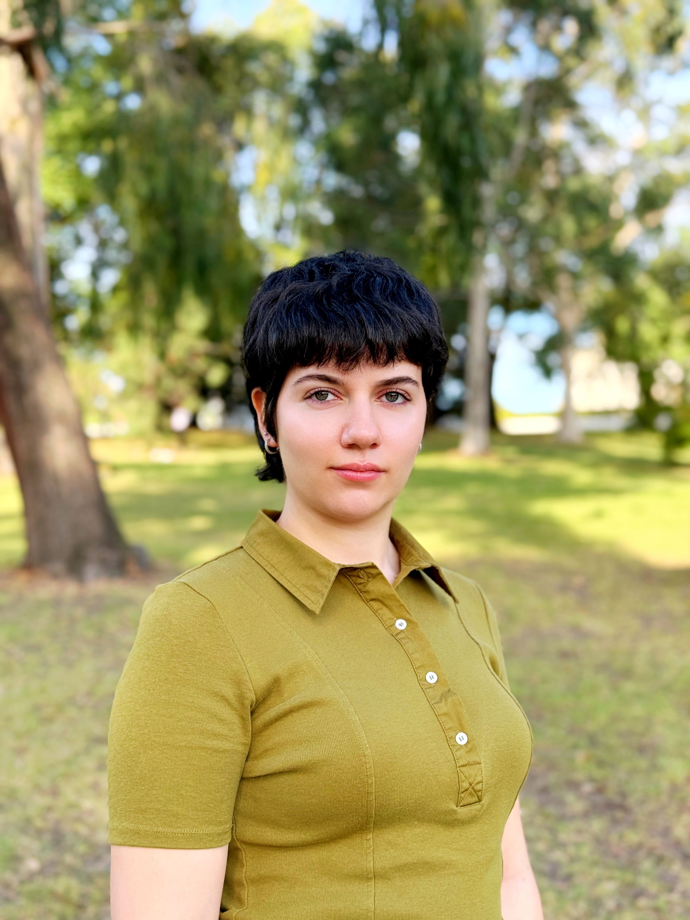 A woman with short black hair, wearing a chartreuse shirt, standing outdoors with gum trees in the background.