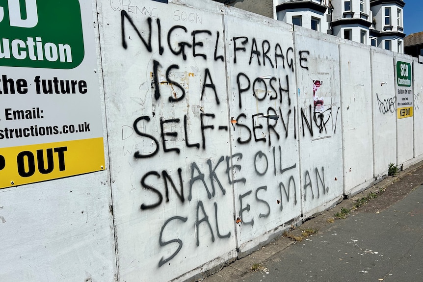 A graffiti message on a white wall that says 'Nigel Farage is a posh self-serving snake oil salesman'
