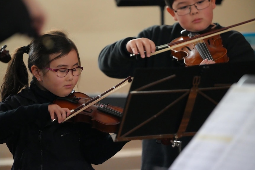 Young boy and young girl both playing violin. Both are wearing black shirts and looking at a music stand.