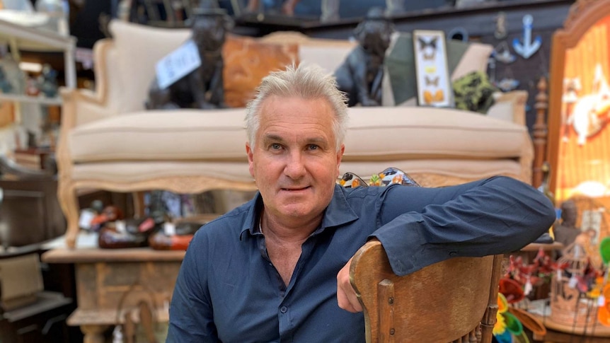 Older man sits on antique chair, with antiques and collectables in the background of his shop.