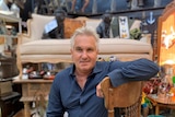 Older man sits on antique chair, with antiques and collectables in the background of his shop.