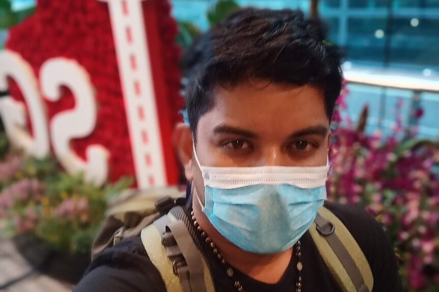 A man with a blue face mask takes a selfie at the airport in front of a red sign.