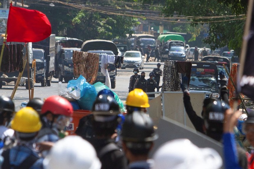 Sarongs are strung up on a wire hanging over a street. Protesters wearing hardhats are on one side, soldiers on the other