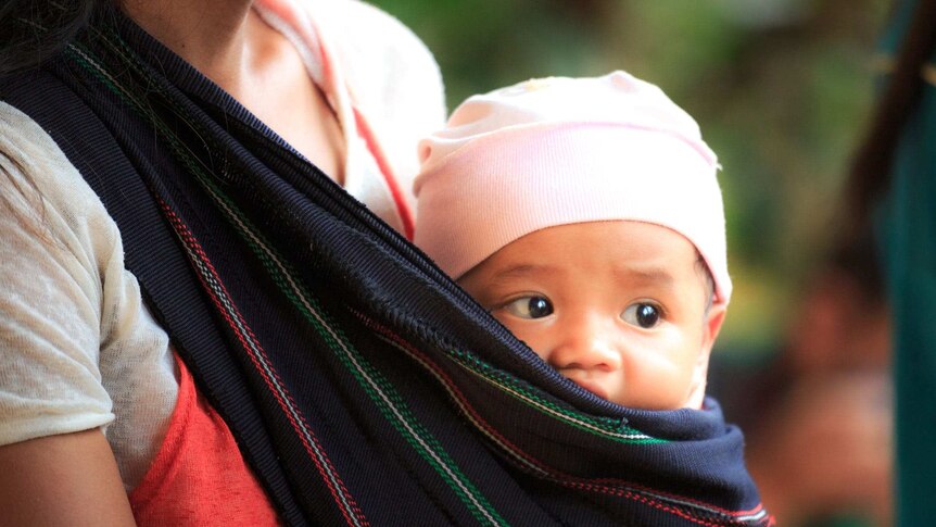 A woman, with her face unseen, has a small baby in a wrap against her body. The baby is looking out from the sling.