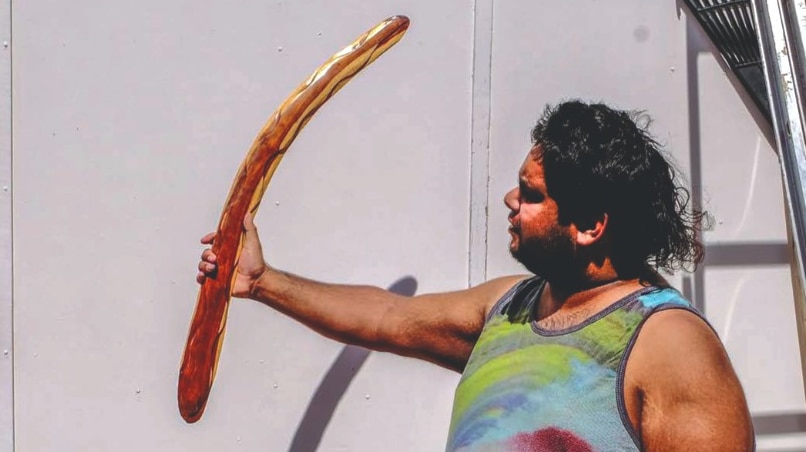 An Indigenous man holds out a long, carved boomerang