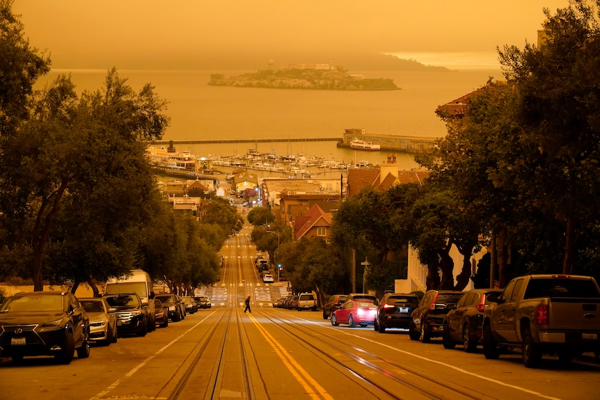 Under an orange sky, a man crosses Hyde Street with Alcatraz Island and Fisherman's Wharf in the background.