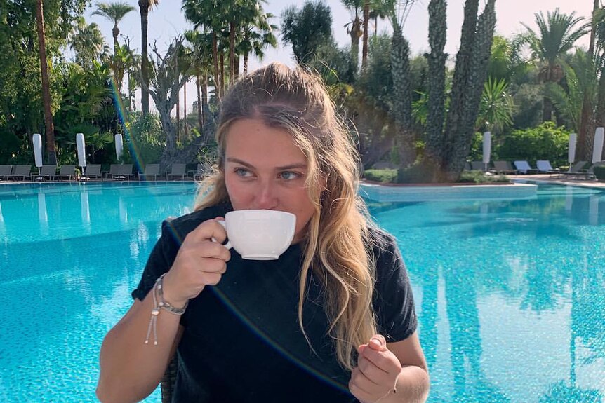 A young blonde woman sips coffee by a lush pool surrounded by date palms