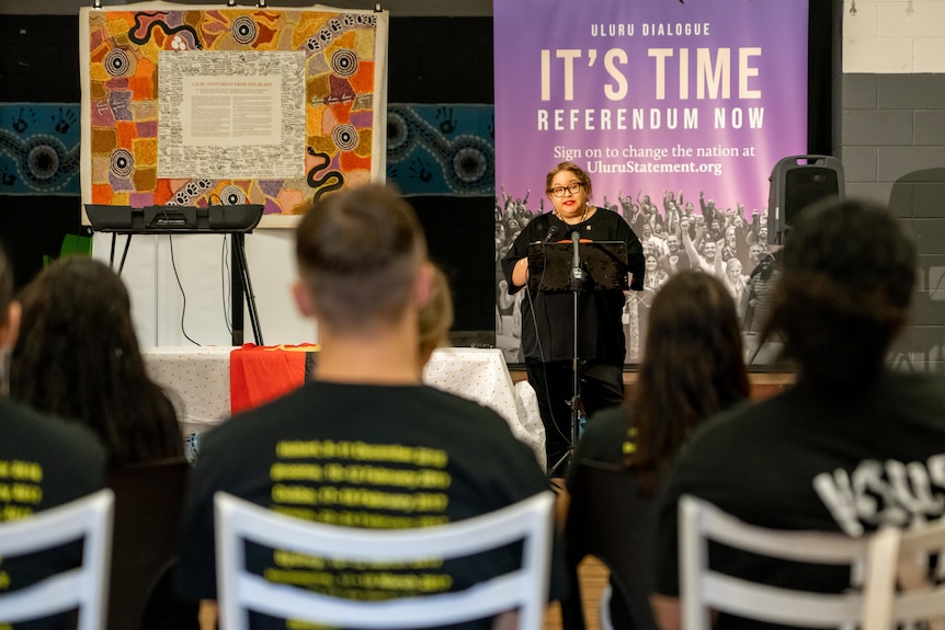 Megan stands at a podium addressing a small crowd, in front of a banner which says "It's Time - Referendum Now".