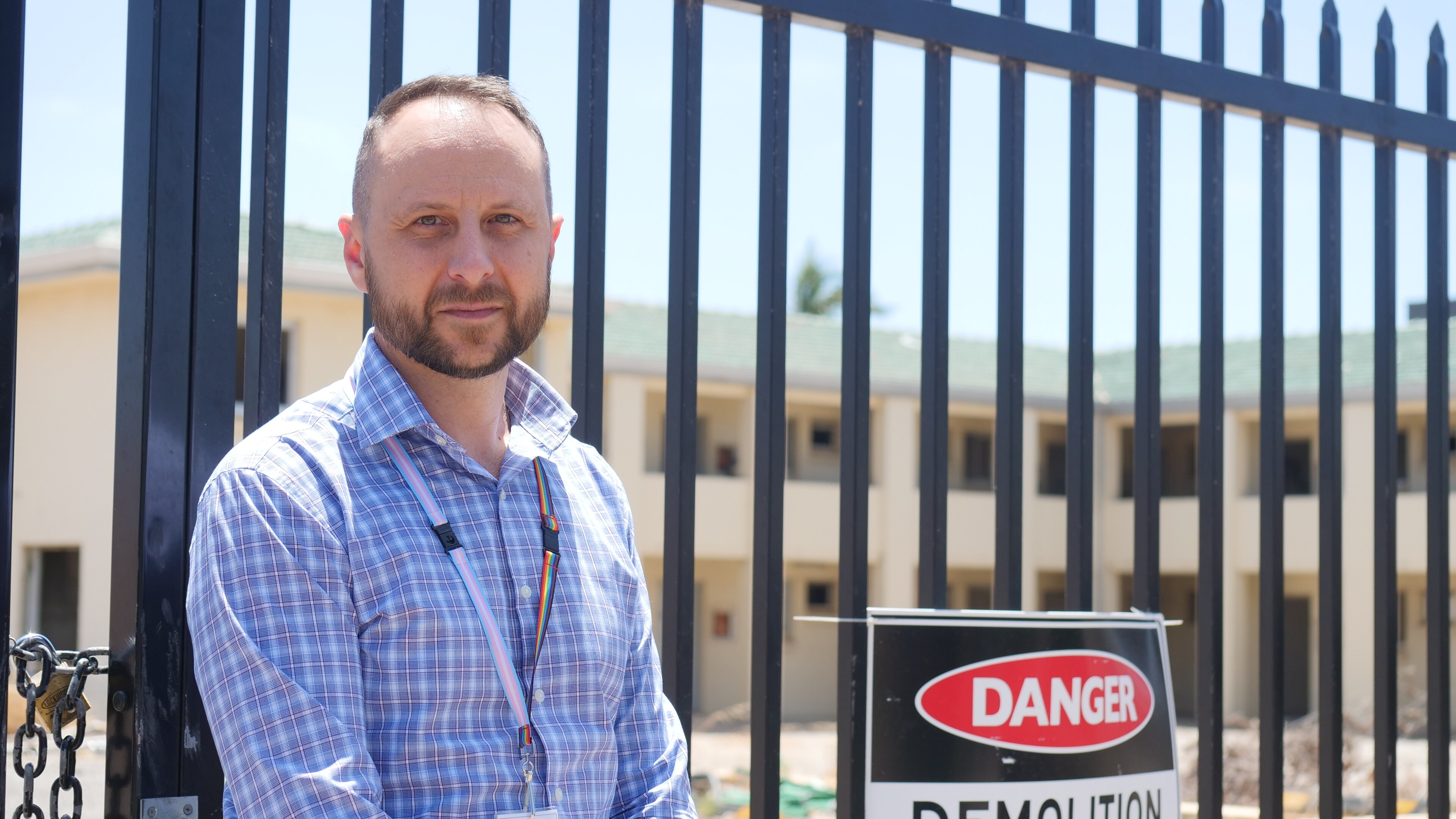 A man in a blue shirt stands next to a fence with a danger sign, looks plainly into camera.