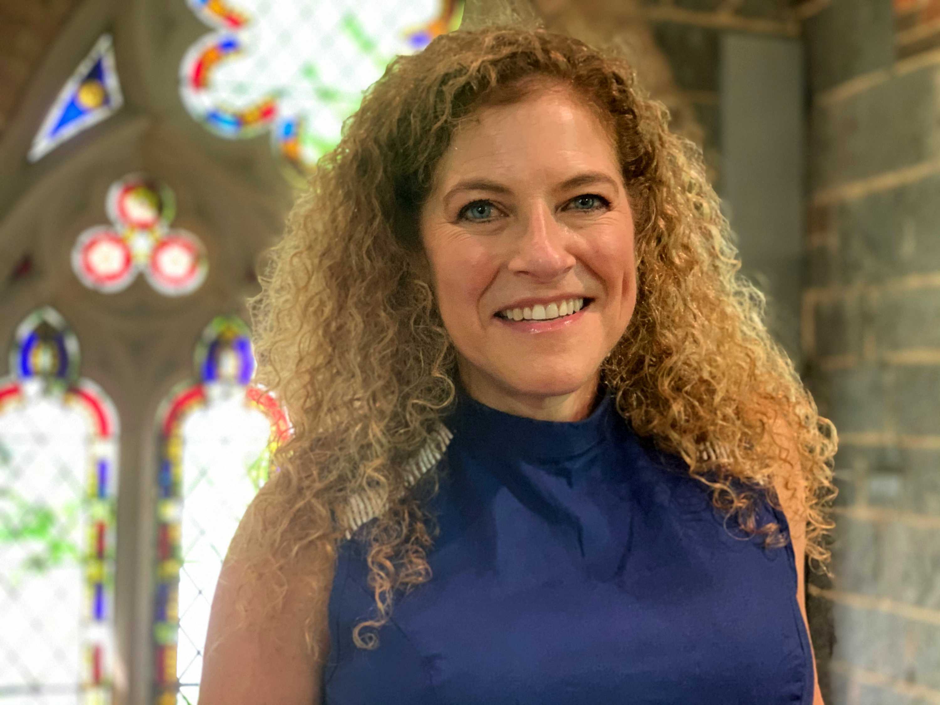 A woman with curly hair smiles as she stands in front of stained glass windows.