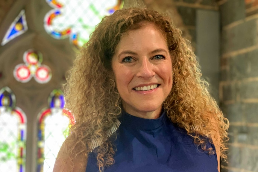 A woman with curly hair smiles as she stands in front of stained glass windows.