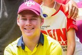 A teenage boy wearing a maroon cap and a high-vis yellow collared shirt kneels smiling at the camera.
