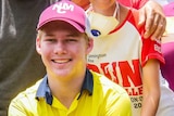 A teenage boy wearing a maroon cap and a high-vis yellow collared shirt kneels smiling at the camera.