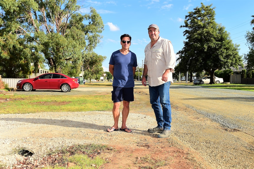Two men standing in a dirt driveway at the edge of a street, with trees in the background.