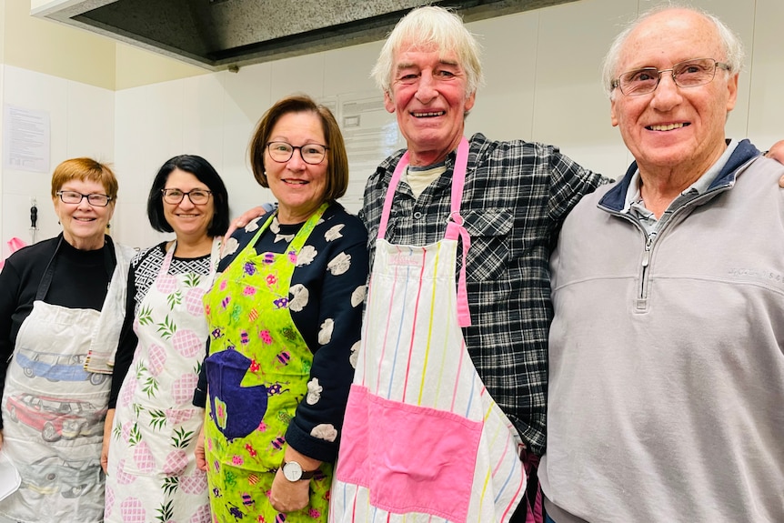 Five people stand wearing colourful aprons in a kitchen and smile