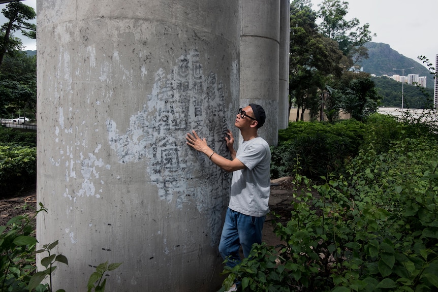 A man in a grey shirt with a black beanie touches his hand to a concrete pillar with fading black calligraphy painted on side.