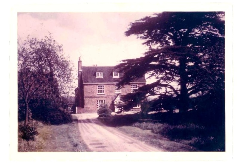 An old sepia photograph of a three-storey wooden cottage surrounded by trees with a long rubble driveway.