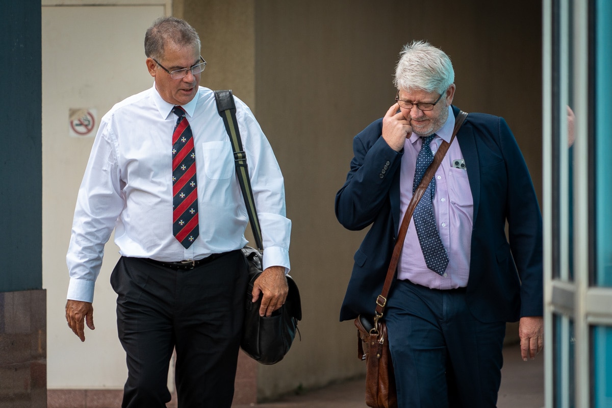 Two middle-aged men in formal attire carry shoulder bags as they walk away from a large building.