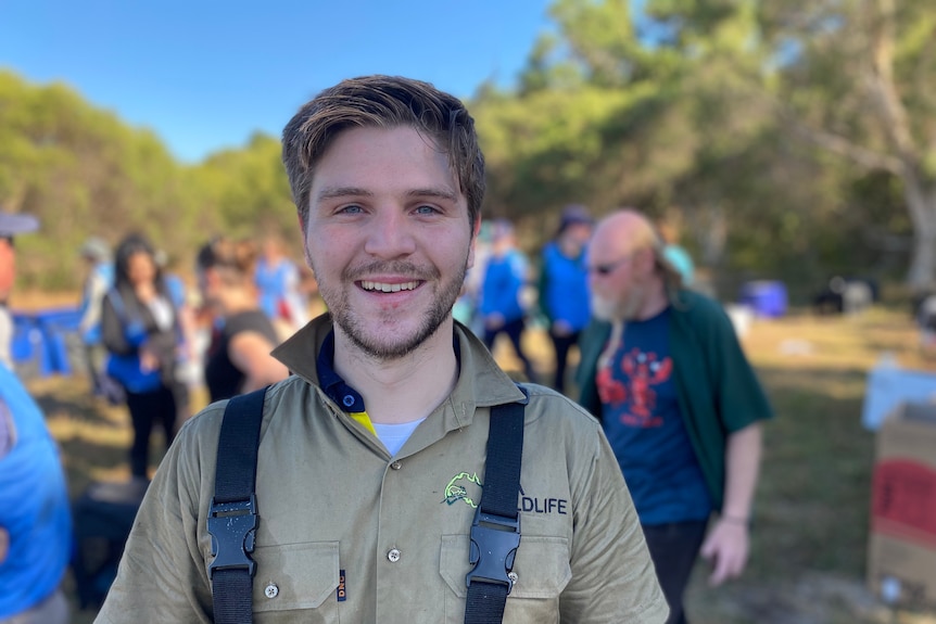 Young man with blonde hair and moustache smiles, with volunteers in the background