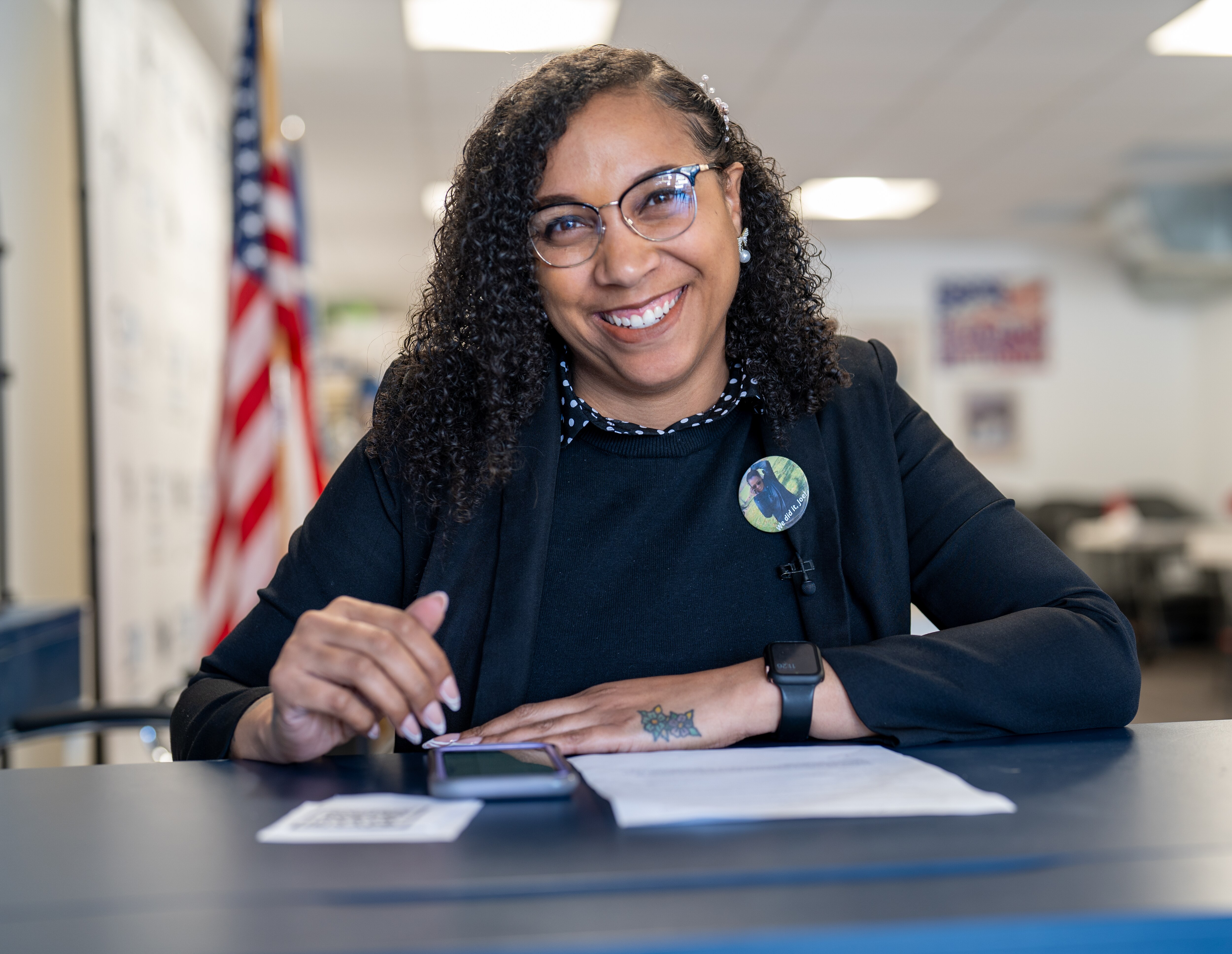 Woman wearing a black top and jacket sitting at a desk with a phone in front of her.