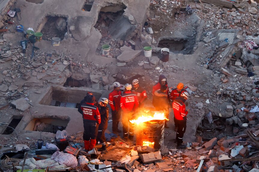 Emergency workers huddle around fire at site of collapsed building.