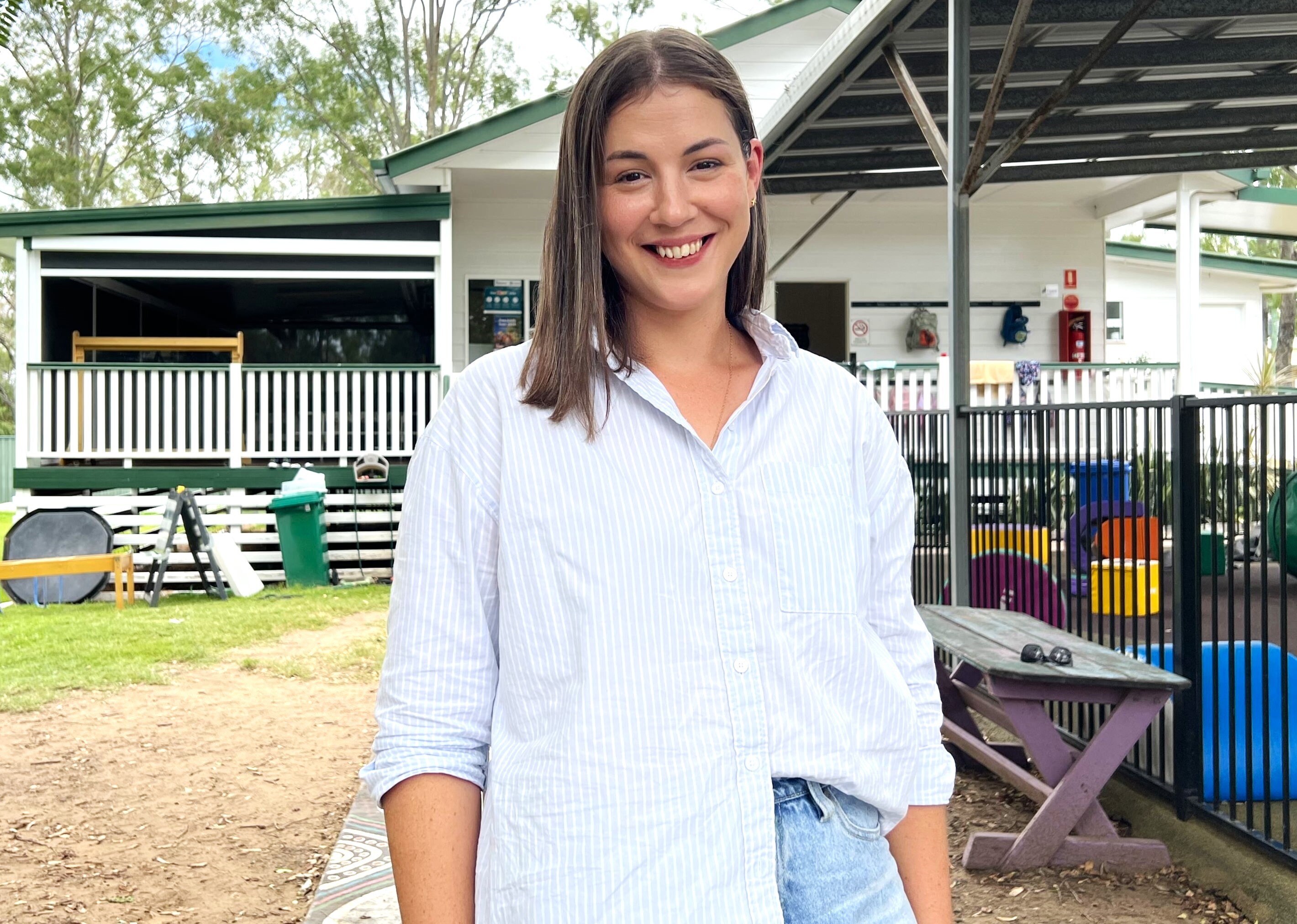 A young woman smiles standing in front of a small kindergarten, there's play equipment behind her