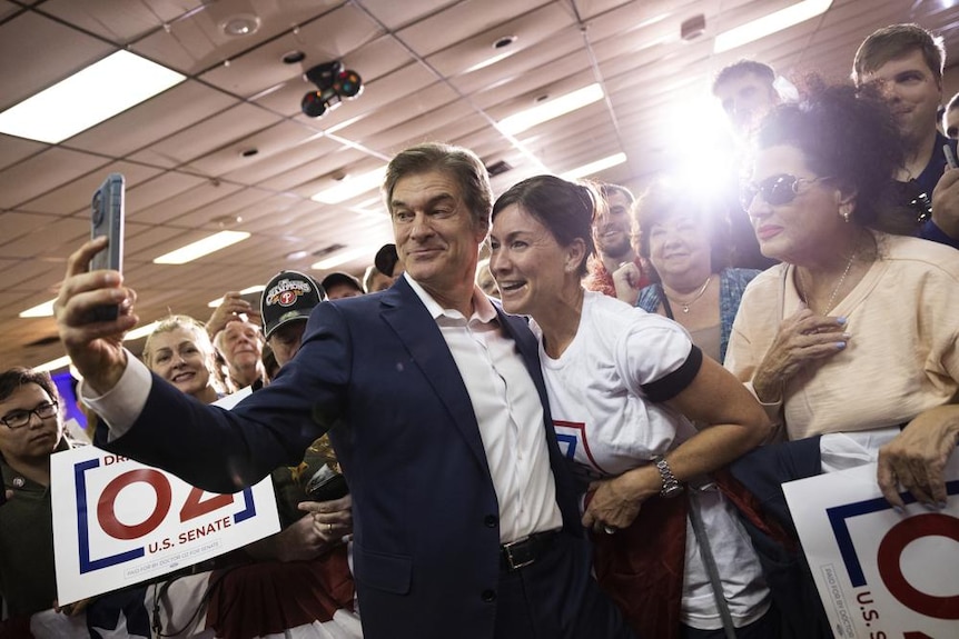 A man, Dr Oz, wearing a navy suit takes a selfie with a woman. Both are surrounded by crowds.