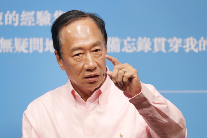 A middle-aged man in pale pink shirt holds his hand up as he gestures with two fingers