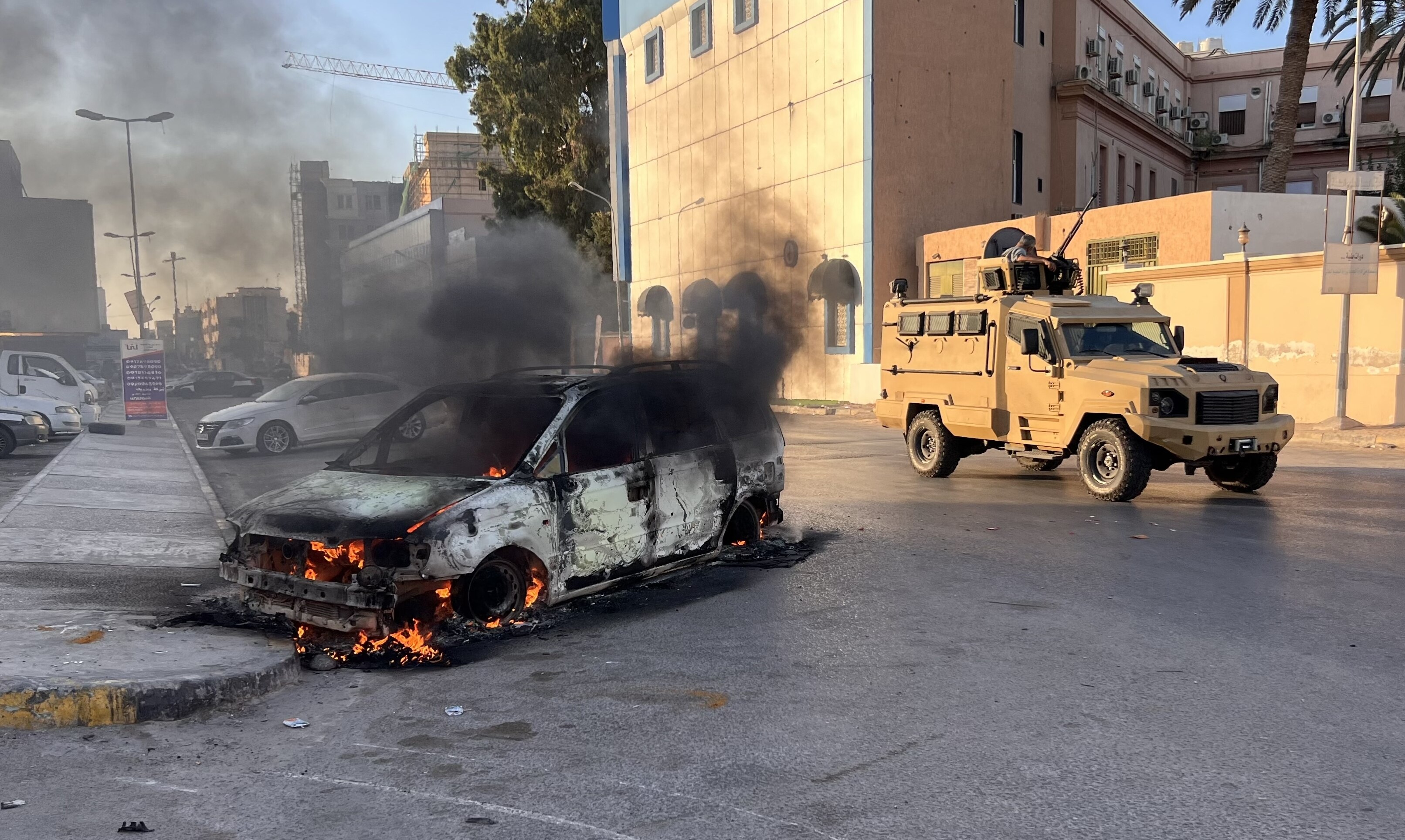 A wrecked vehicle burns at the street as a military vehicle drives past,