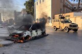 A wrecked vehicle burns at the street as a military vehicle drives past,