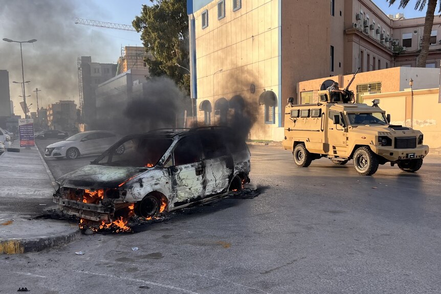 A wrecked vehicle burns at the street as a military vehicle drives past,