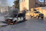 A wrecked vehicle burns at the street as a military vehicle drives past,