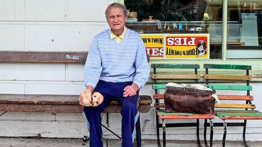 A man sits outside a milk bar holding the skull of a two headed calf foetus