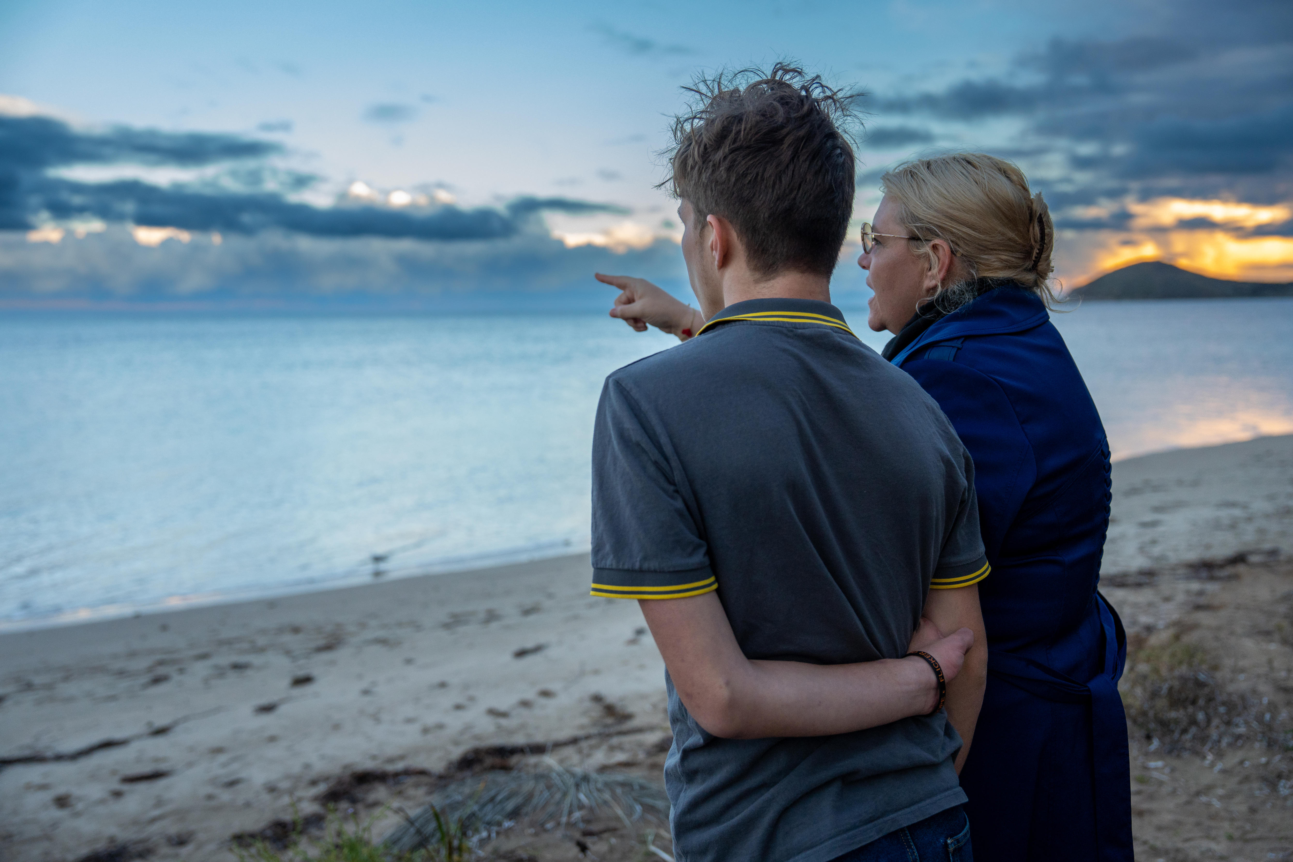 A son and mother stare out to an ocean, the mother pointing to the horizon