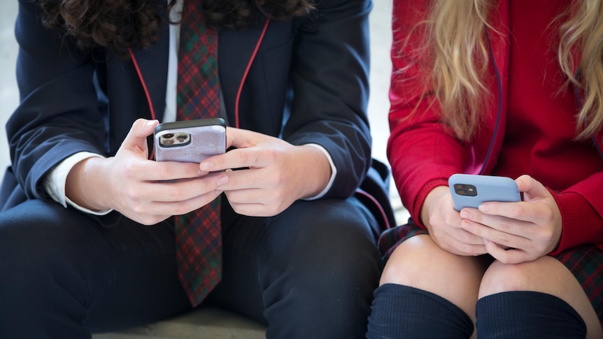 A generic photo of two teenage schoolkids sitting side by side, using their phones.
