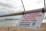 A beach closed sign in front of a beach with wharf in the background.