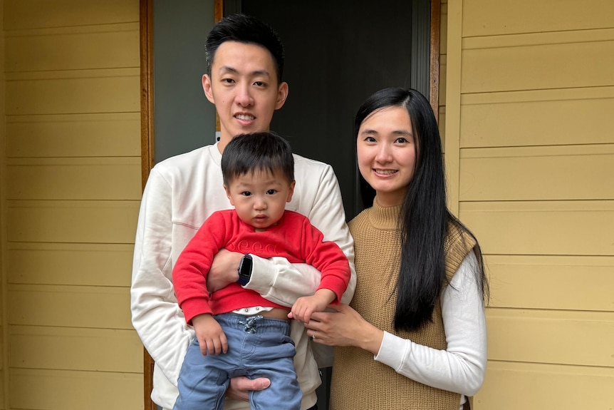 Adrian holds his young son and stands next to his wife as they smile for a picture at their front door.