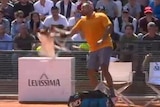 Wearing an orange shirt, Nick Kyrgios hurls a small white stool onto the clay court while the crowd watches on.