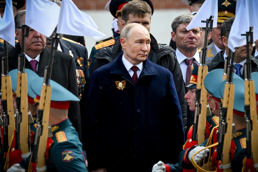 A man in a dark coat stands among a military parade