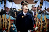 A man in a dark coat stands among a military parade