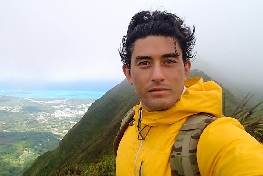 Nathan Crumpton takes a selfie high up in the hills with a view to mountains and water behind him.