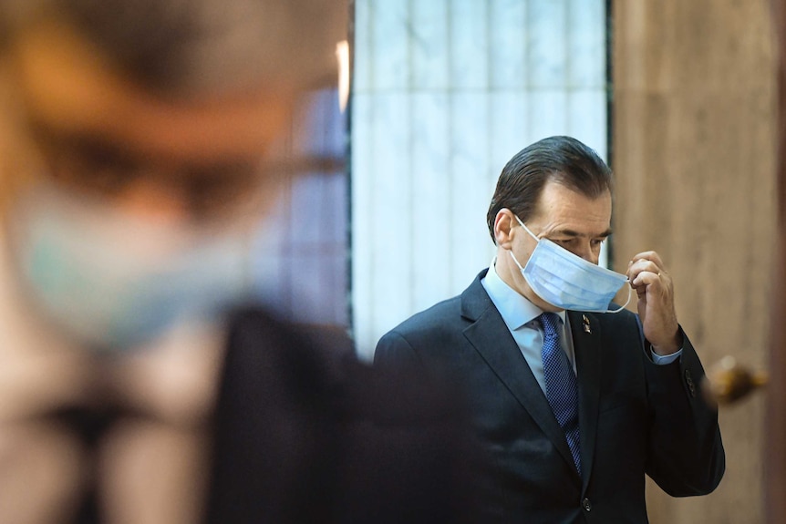 a man wearing a suit touches the face mask he is wearing