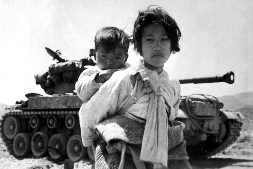 Black and white photo Korean girl with baby on her back stands in front of a tanker looking somber
