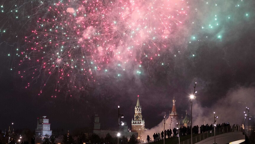 The Kremlin is illuminated as red and green fireworks explode in the sky above it.