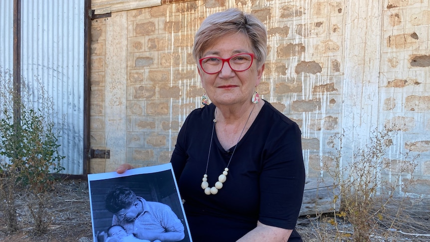 A woman holding a photo of her father, who is holding a baby, sitting on a log in front of a brick wall. 