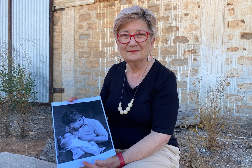 A woman holding a photo of her father, who is holding a baby, sitting on a log in front of a brick wall. 