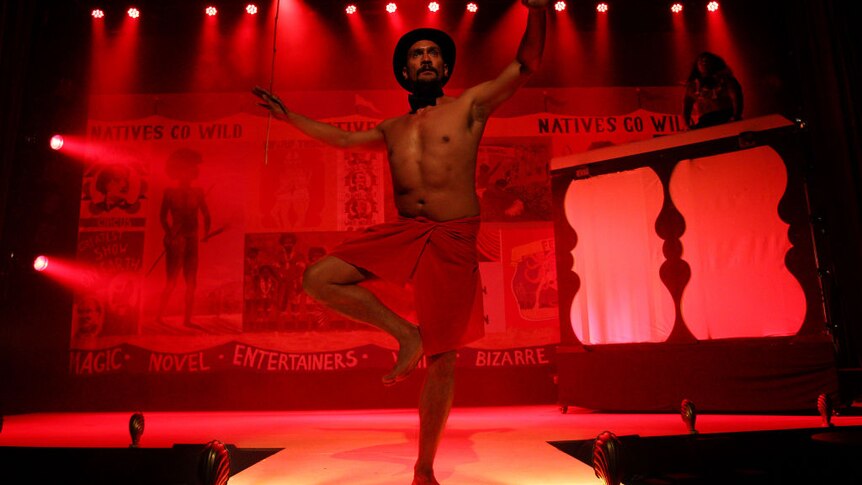 Red-lit cabaret stage. Bare-torso performer stands on one leg with other bent so foot on knee, balancing, wearing red lap lap.