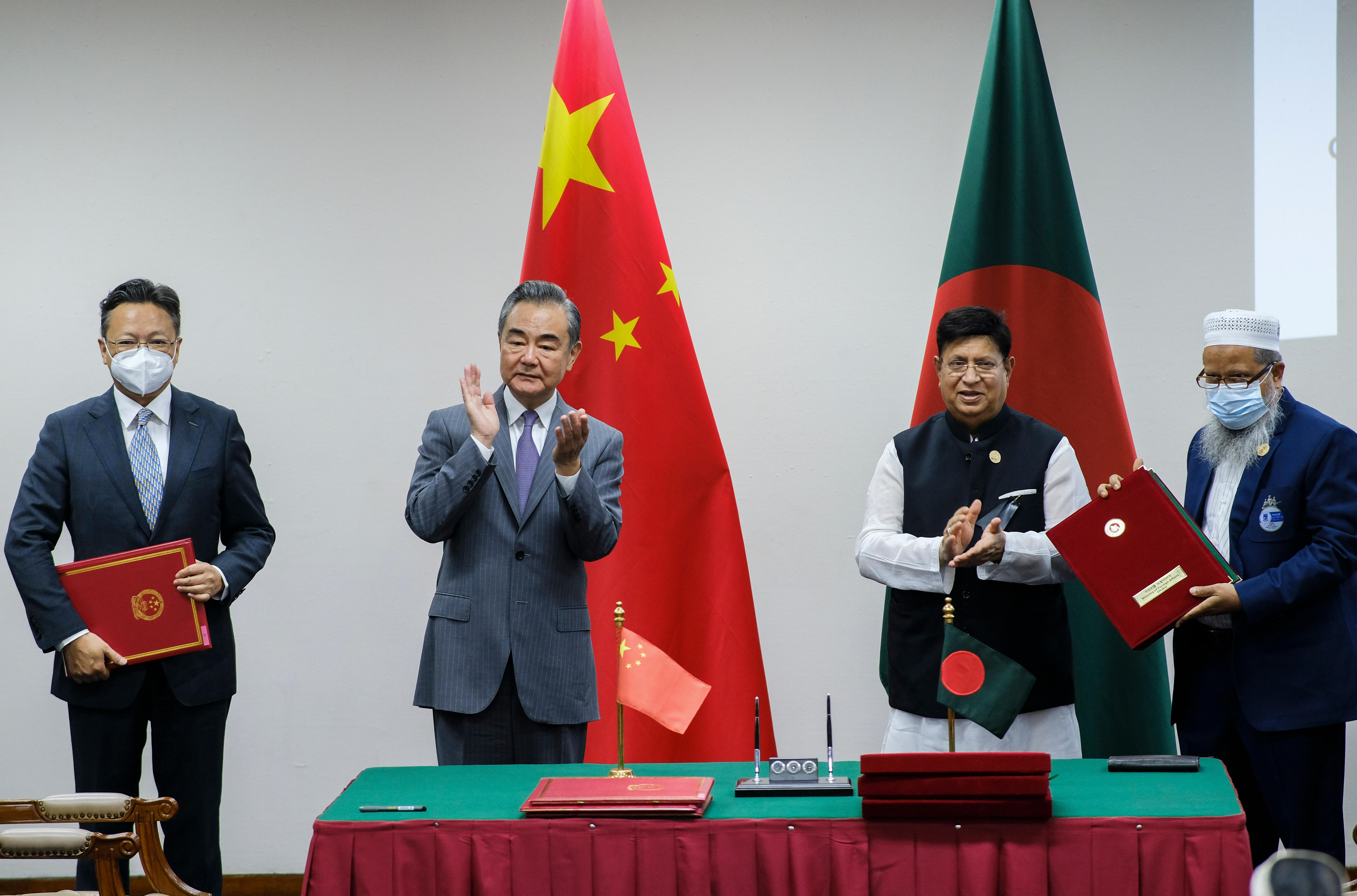 Four suited men stand behind a table, in front of the Chinese and Bangladesh flags, applauding