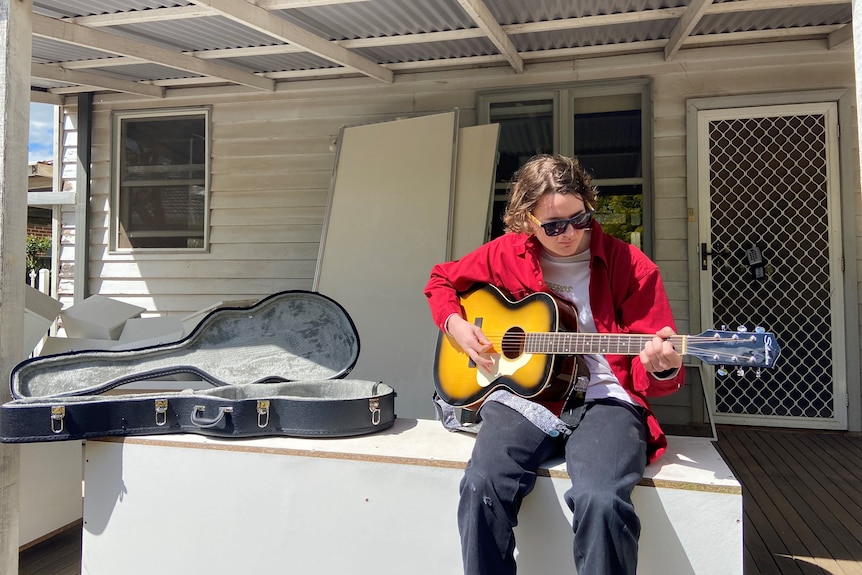 A photo of a a young guy with a guitar sitting on furniture on a verandah.